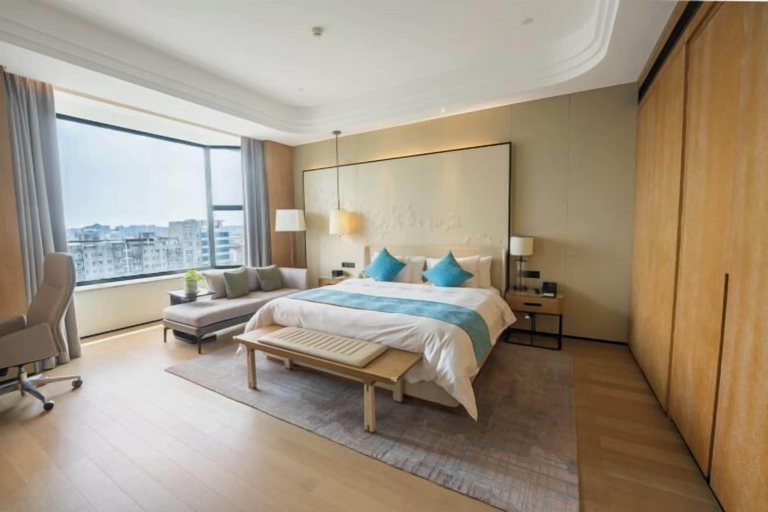 A modern hotel room with a large bed, a desk, a chair, and a view of the city.