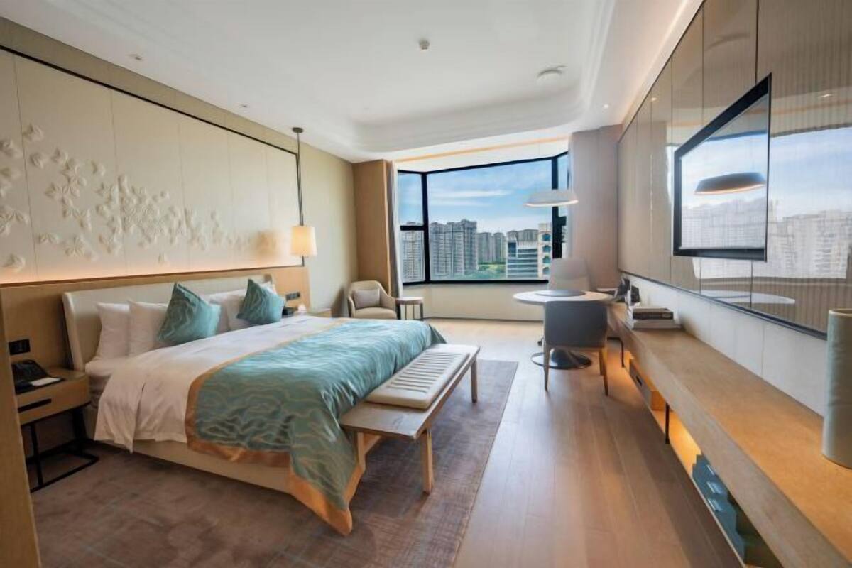 A modern hotel room with a large bed, a desk, a chair, a small table, and a view of the cityscape through large windows.