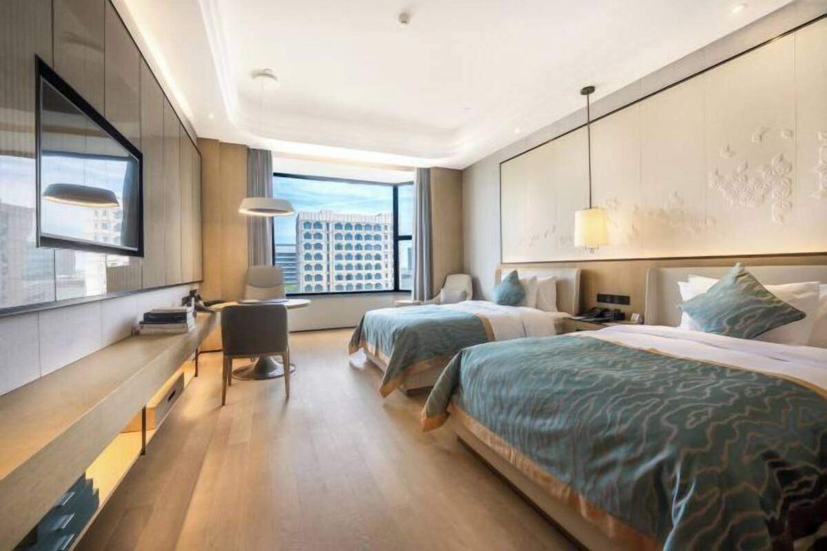 A hotel room with two beds, a desk, a chair, a TV, and a large window with a city view.