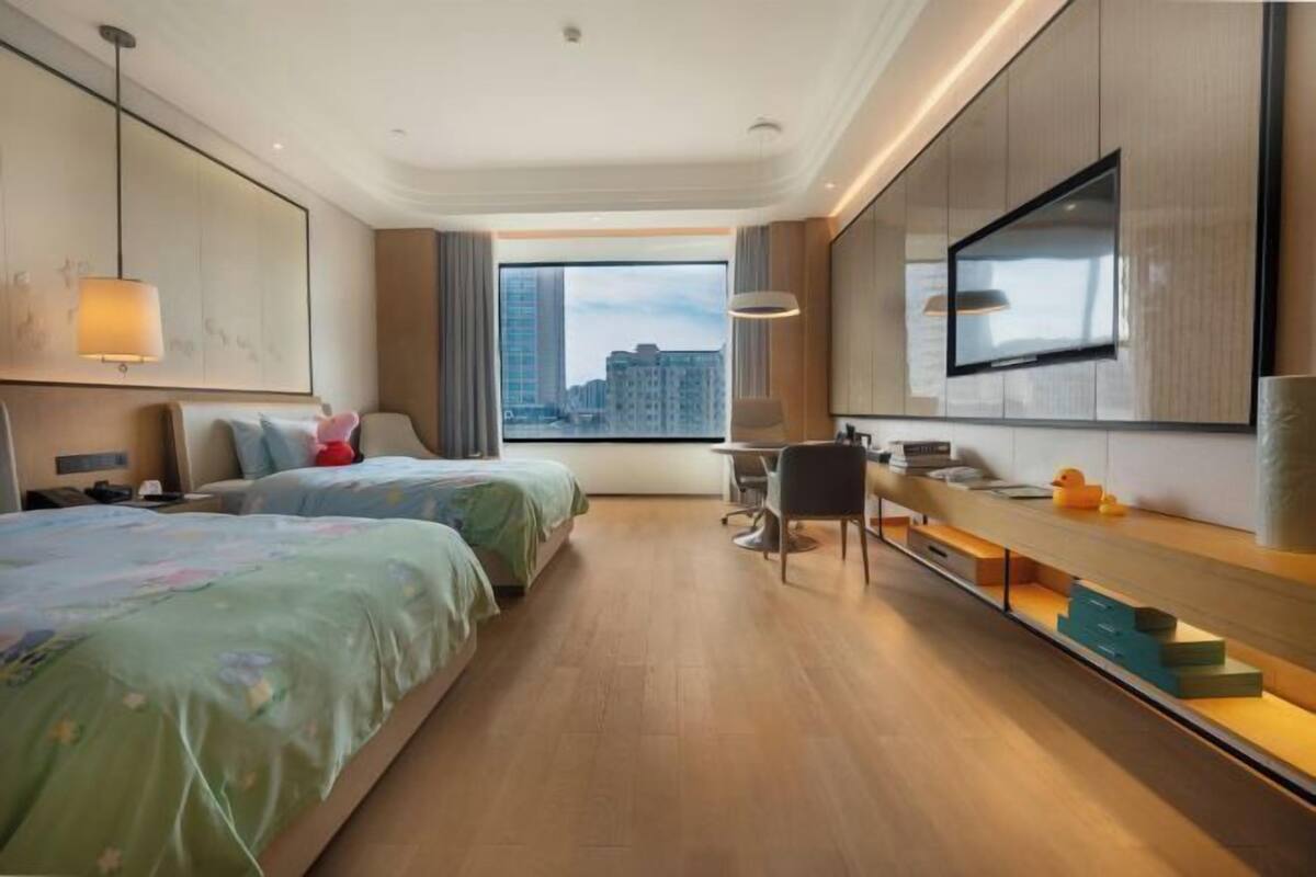 A hotel room with two beds, a desk, a chair, a TV, and a large window with a city view.