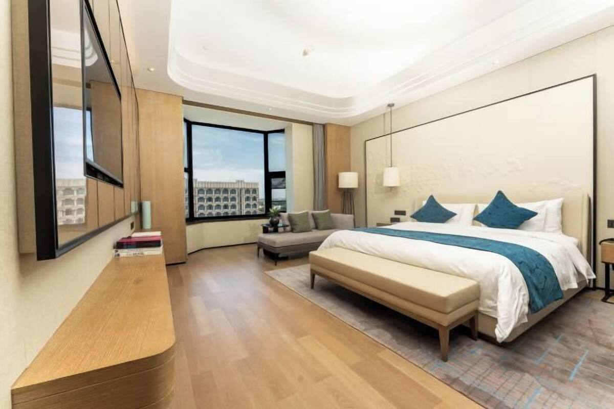 A modern hotel room with a large bed, a bench, a desk, and a view of the city.