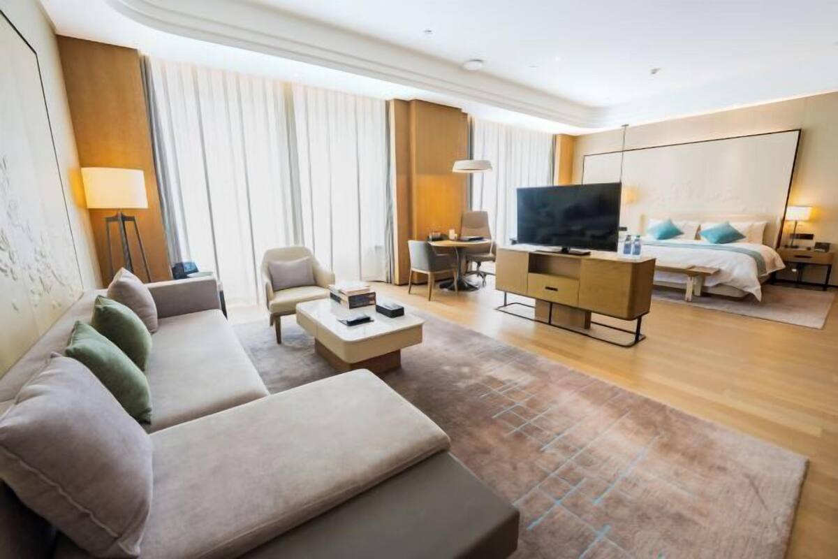 A hotel room with a sofa, a TV, a desk, a bed, and a large window.