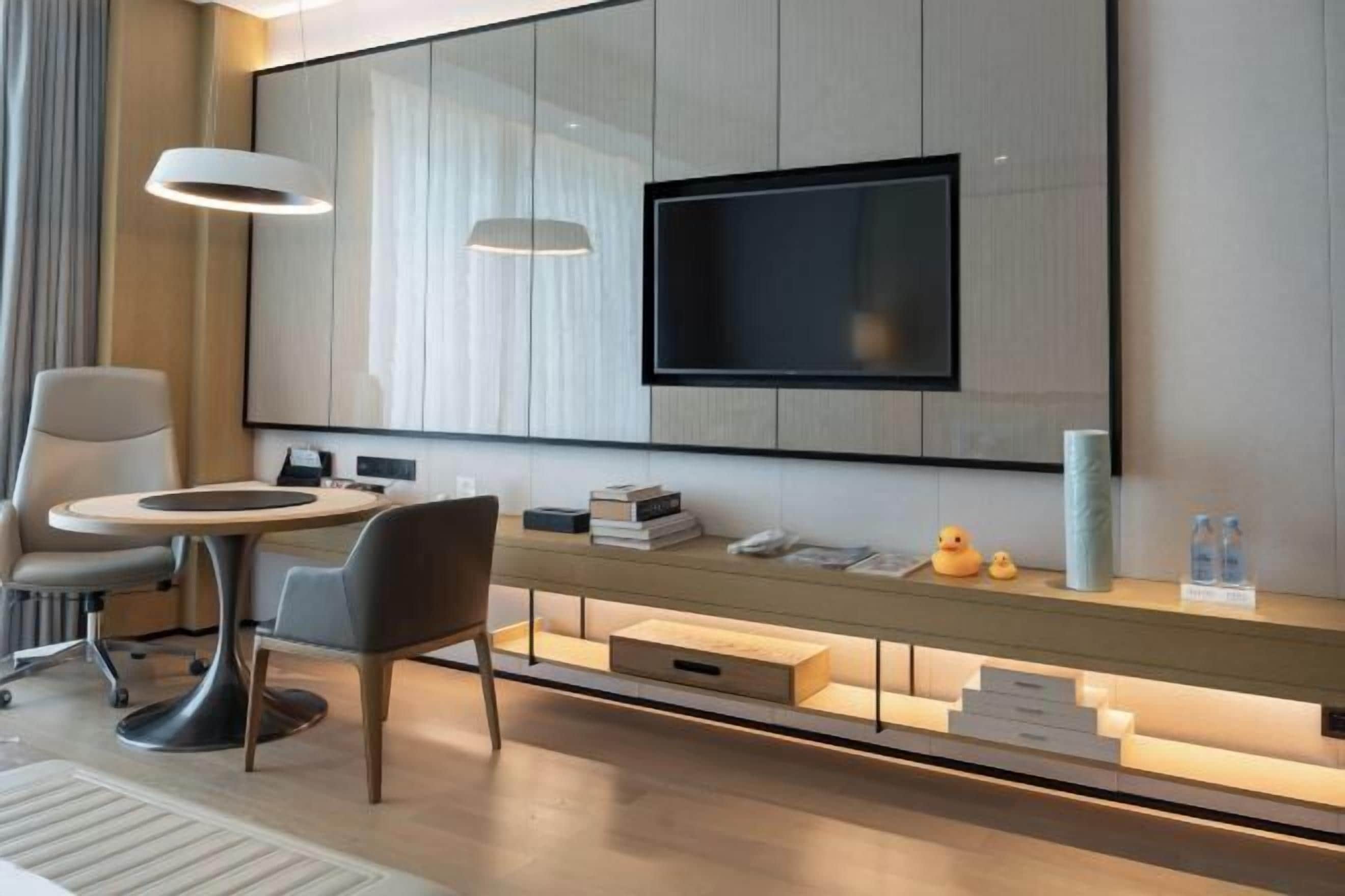 A modern hotel room with a flat-screen TV mounted on a wall, a round dining table with chairs, a built-in bench with storage drawers, and a large window.