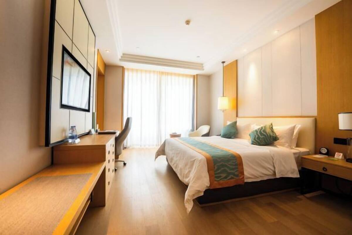 A hotel room with a bed, bedside table, desk, chair, and a large window with curtains.