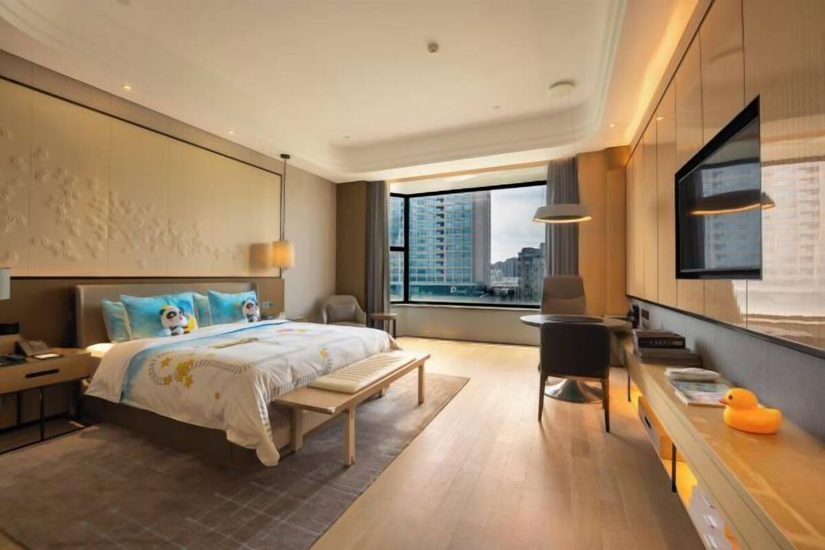 A modern hotel room with a large bed, bedside table, and a desk with a chair.