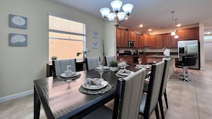 A dining area with a dark wooden table set for four, surrounded by chairs. There is a kitchen in the background with wooden cabinets and stainless steel appliances.