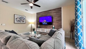 A living room with a ceiling fan, a flat-screen TV mounted on a wooden wall, a glass coffee table, and grey sofas.