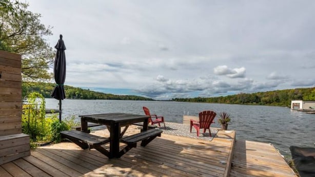 Una terraza de madera con una mesa de picnic y sillas rojas, con vista a un lago bajo un cielo nublado.