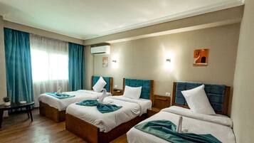 Quarto de hotel com três camas, uma mesa pequena e uma janela com cortinas.