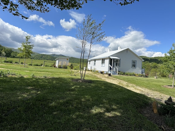 A white house with a porch, surrounded by a grassy field and trees, with mountains in the background.