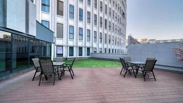 A rooftop terrace with a table and chairs, a building in the background, and a small grassy area.