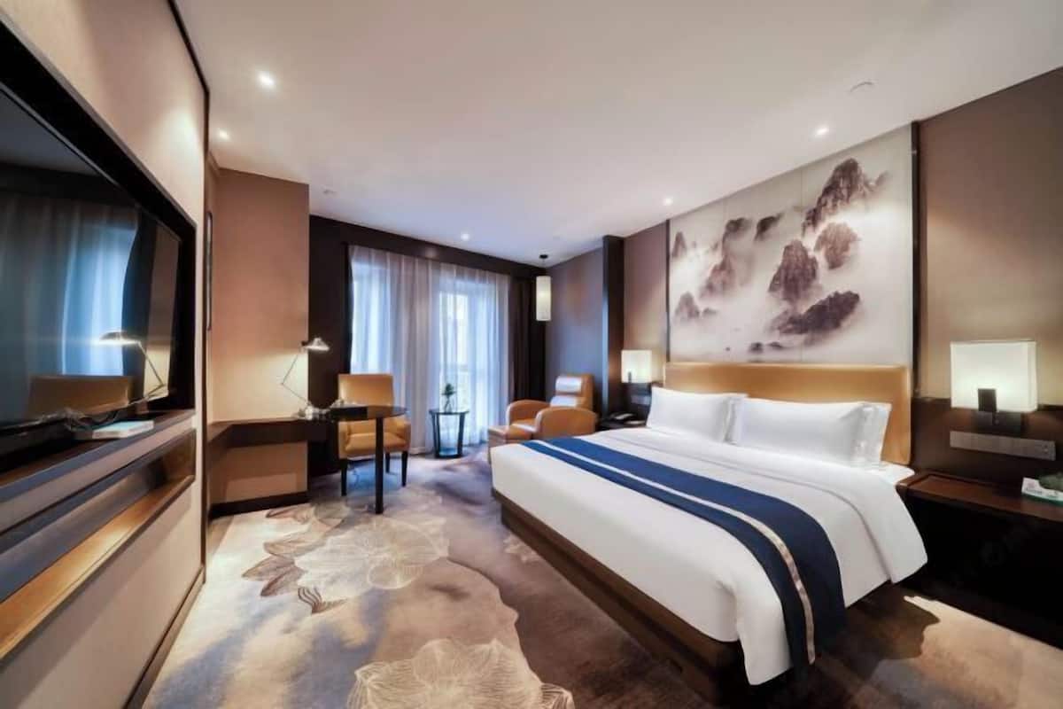 A modern hotel room with a large bed, a desk, a chair, and a large painting on the wall.