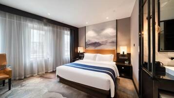 A modern hotel room with a large bed, a chair, a desk, and a large window with sheer curtains.