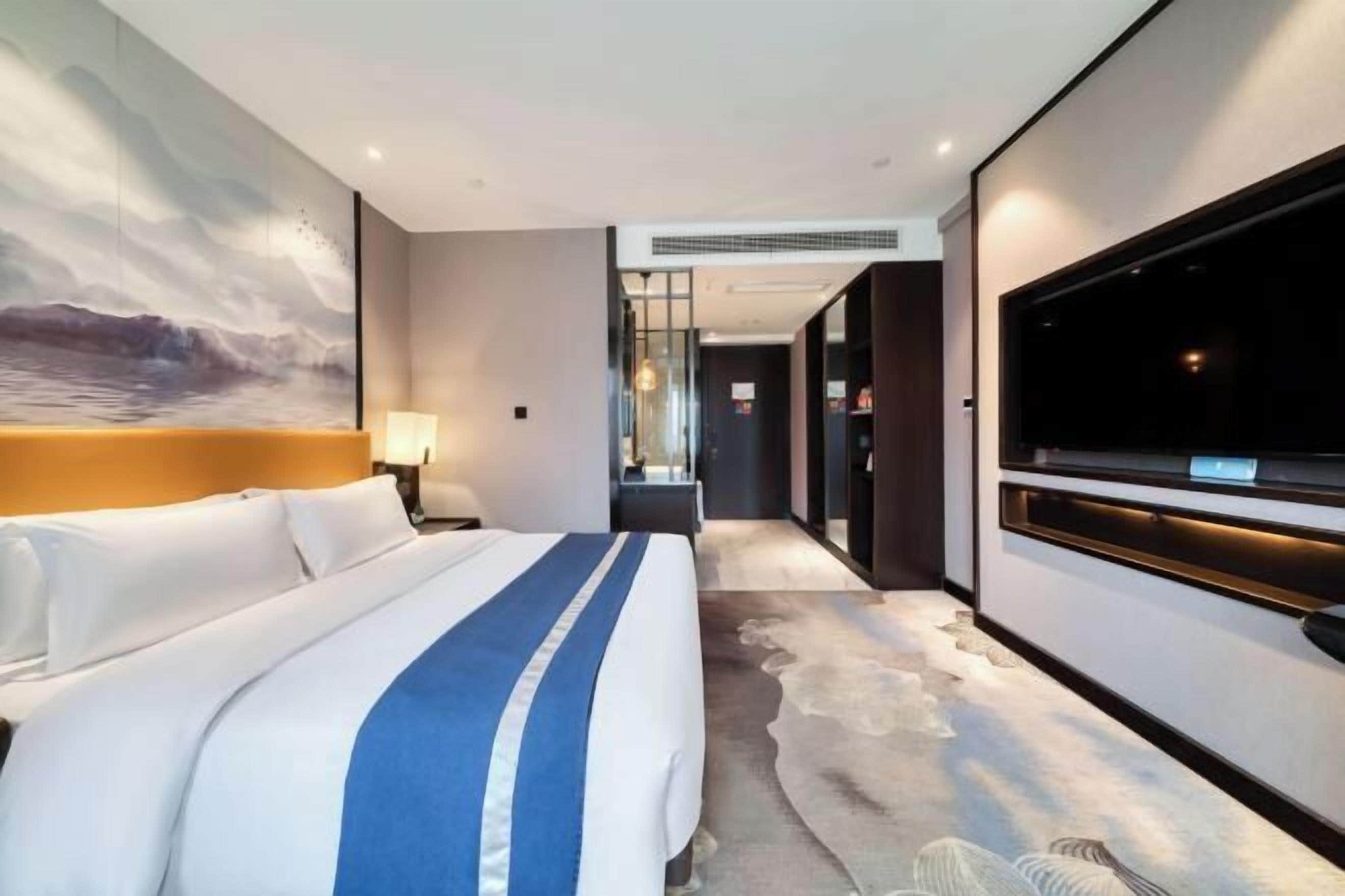 A modern hotel room with a large bed, a flat-screen TV mounted on the wall, and a view of the ocean through the window.
