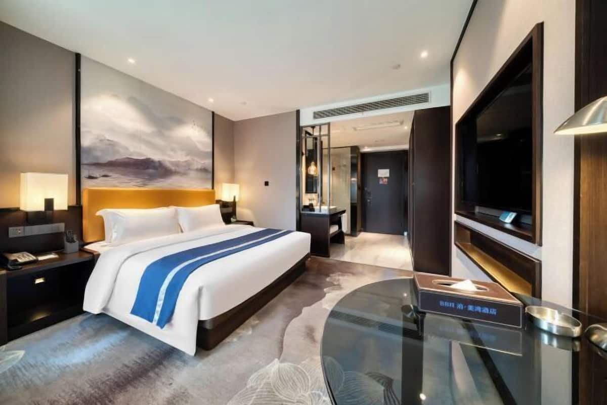 A modern hotel room with a large bed, a glass coffee table, a flat-screen TV, and a large painting on the wall.