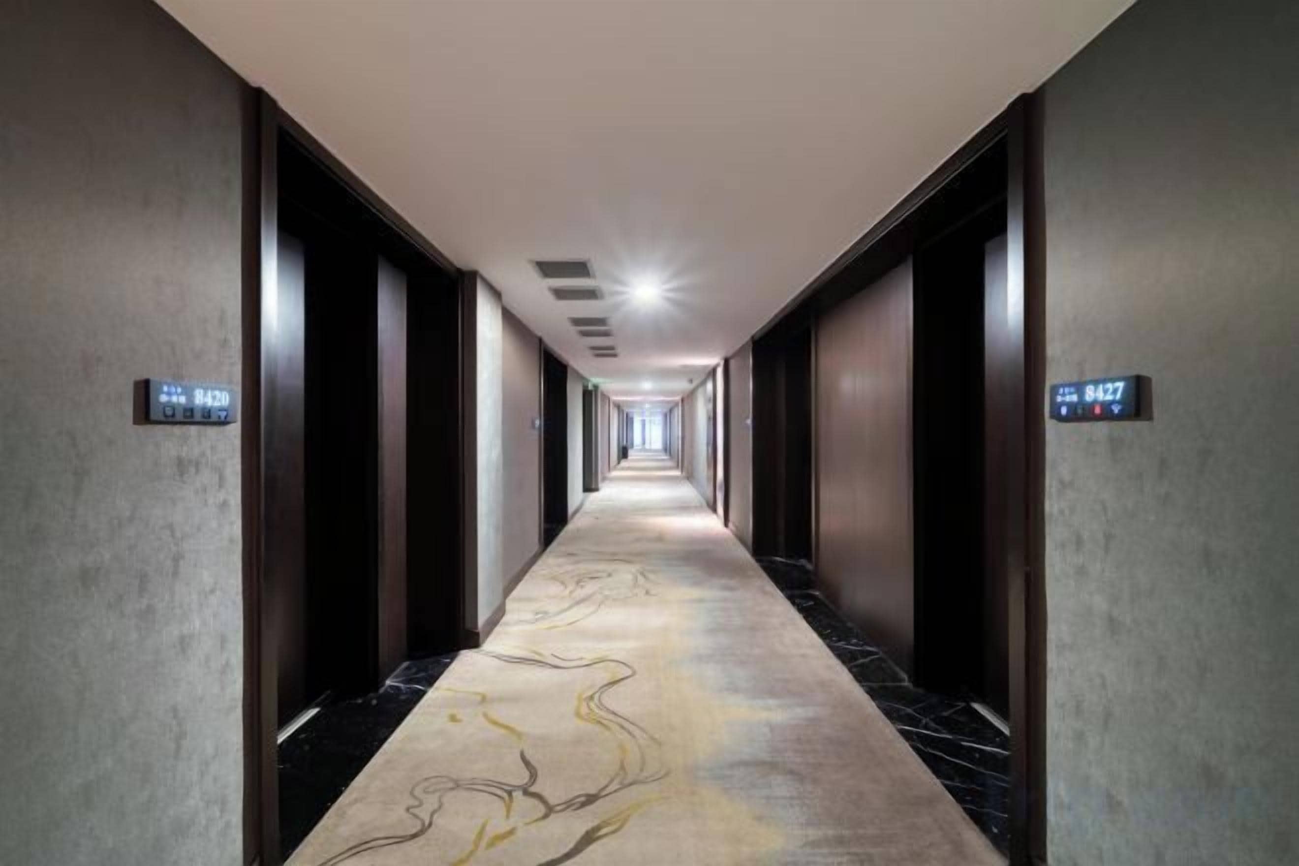 A long, narrow hallway with a patterned carpet, doors on either side, and a digital clock displaying the time.