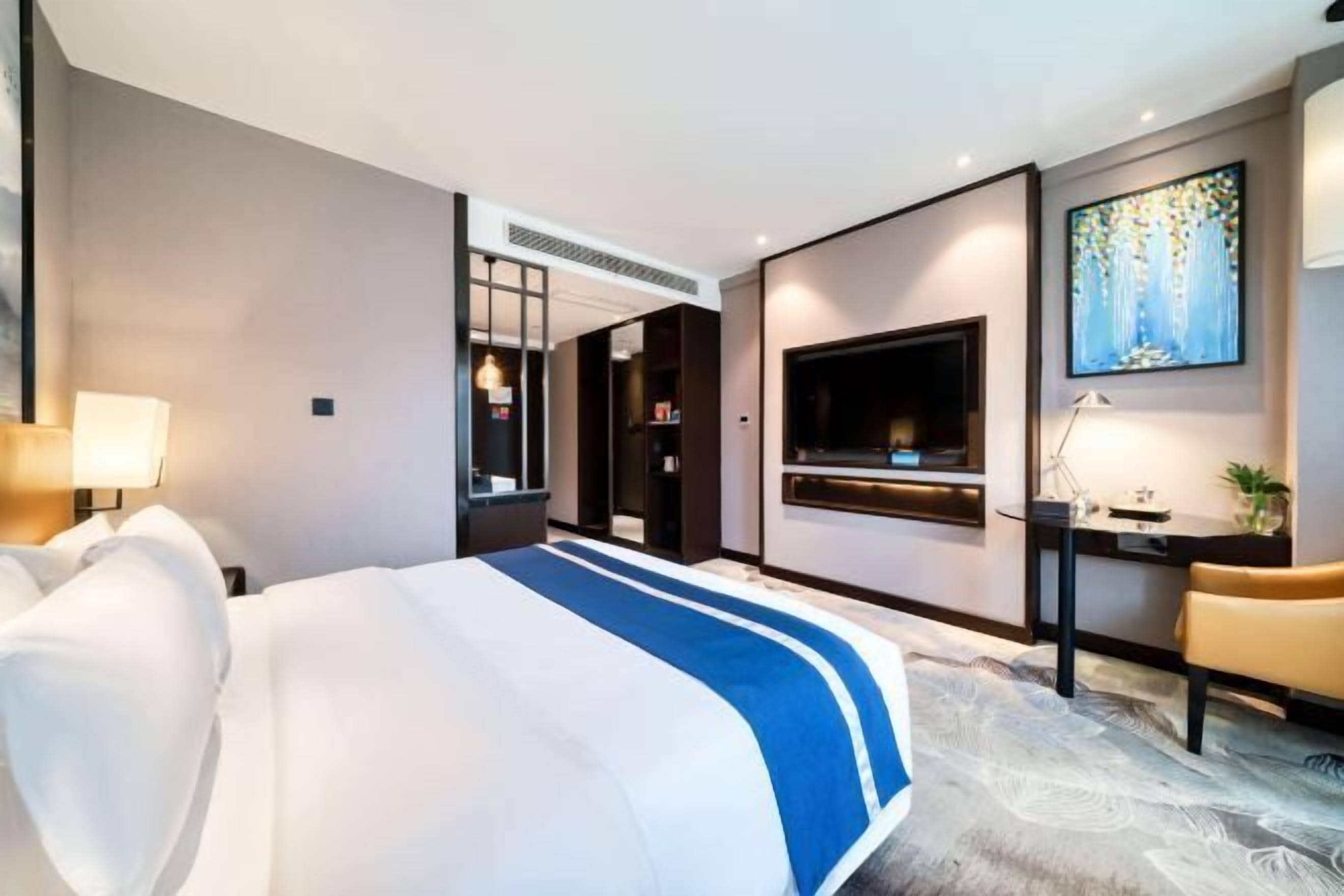 Ein Hotelzimmer mit einem großen Bett, einem an der Wand befestigten Fernseher, einem Schreibtisch mit Stuhl und einem Wandbild.