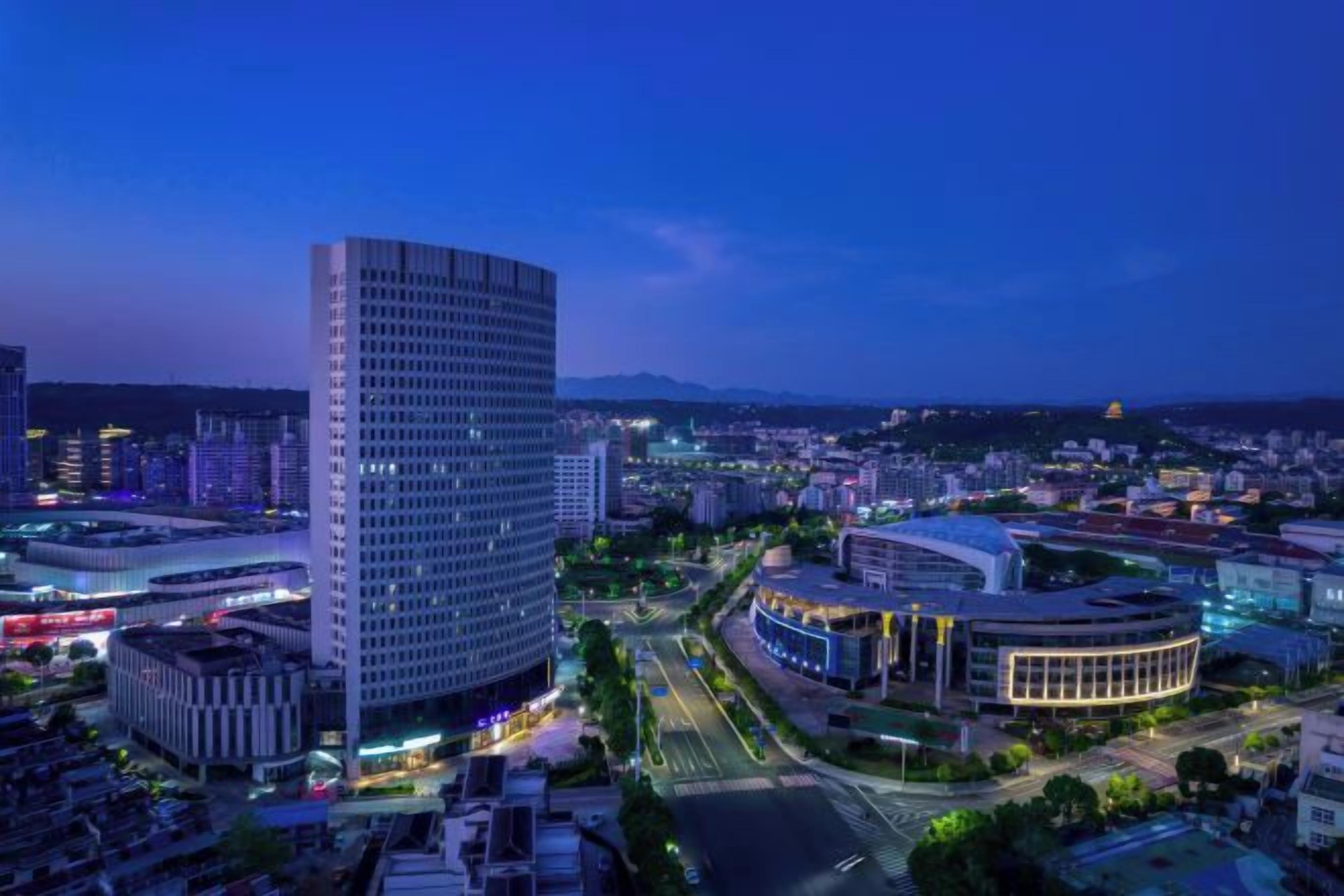 Un paisaje urbano al anochecer con edificios modernos, incluyendo una estructura curva destacada y un edificio rectangular alto.