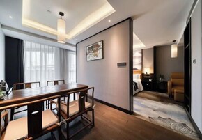 Una habitación de hotel moderna con una mesa de comedor de madera, un sofá y un ventanal con cortinas.