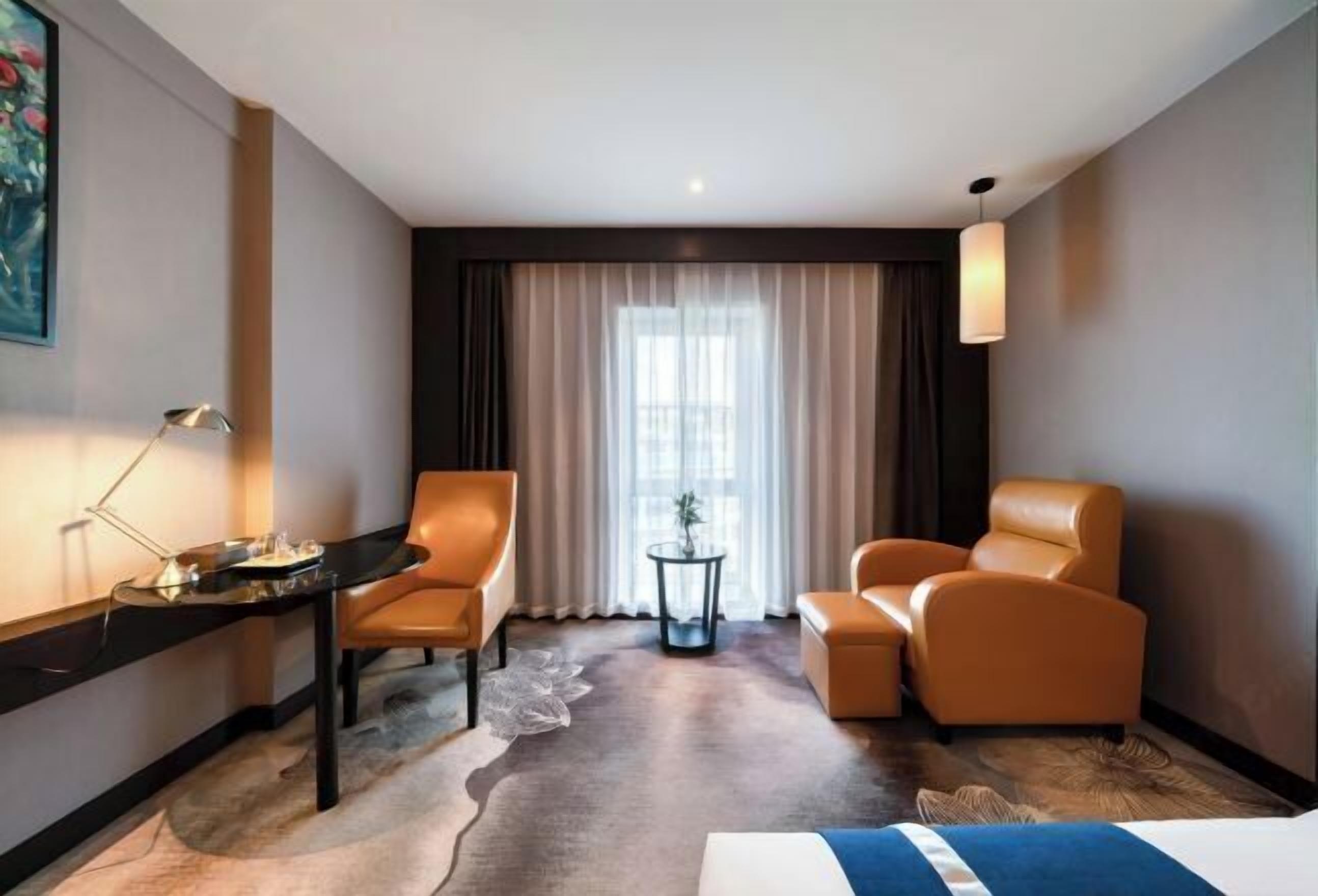 Habitación de hotel con cama, dos sillones, un escritorio con lámpara, una mesita con planta y un ventanal con cortinas.