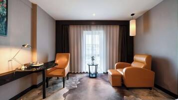 Habitación de hotel con cama, dos sillones, un escritorio con lámpara, una mesita con planta y un ventanal con cortinas.