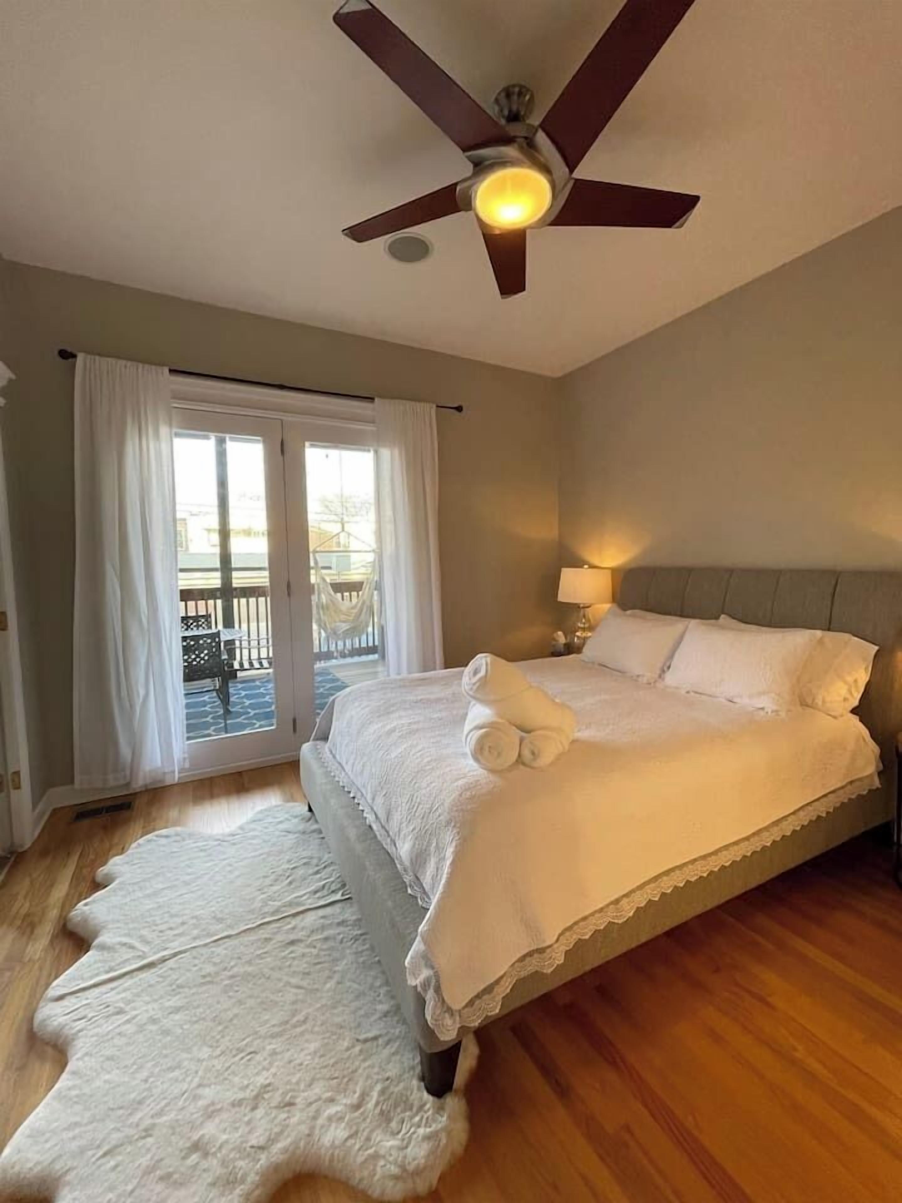 A bedroom with a large bed, a ceiling fan, a nightstand with a lamp, and a view of a balcony through the glass door.