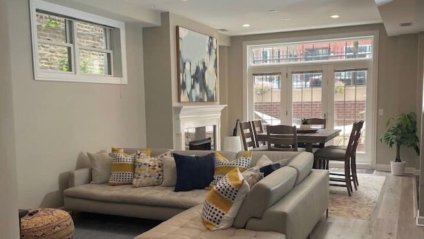 A modern living room with a sectional sofa, a fireplace, and a dining area with a view through large windows. - Ukrainian Village 3BR/3BA • Private Patio, Parking (Chicago)
