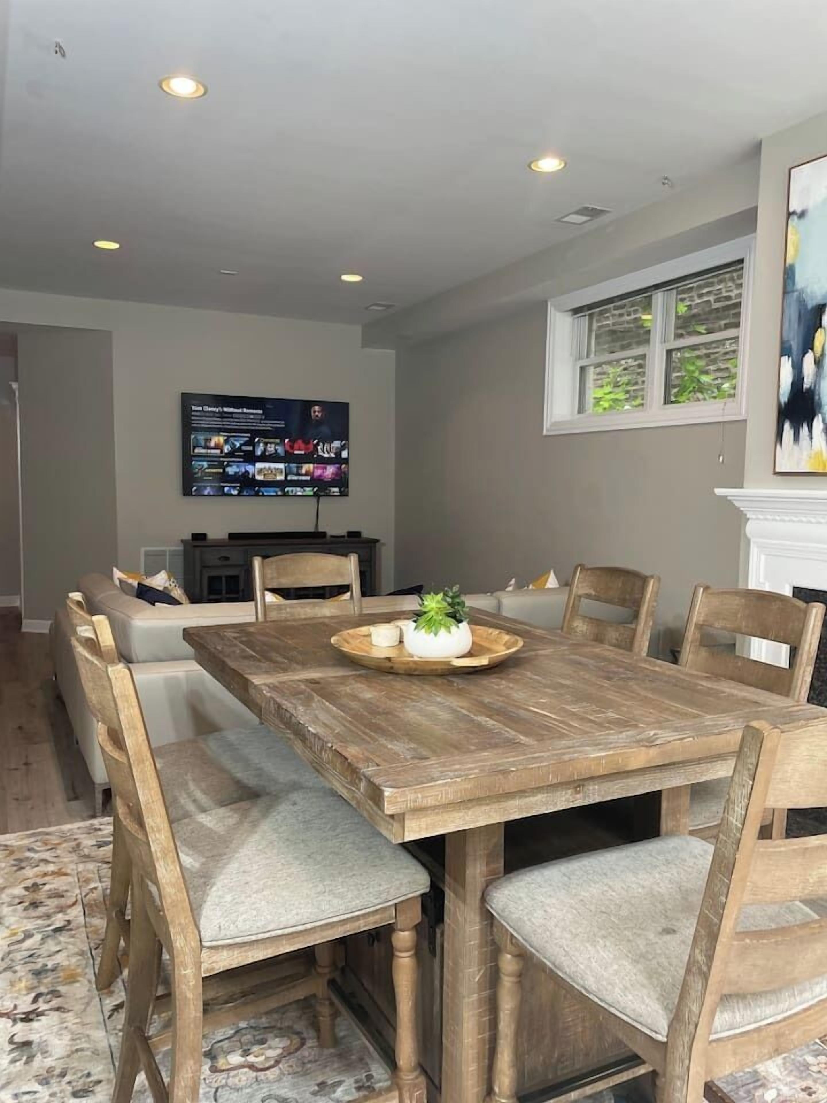 Une salle à manger avec une table en bois et six chaises, un téléviseur à écran plat fixé au mur et une fenêtre donnant sur un paysage verdoyant.