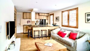 A modern kitchen with wooden cabinets, a central island with stools, a dining area with a sofa and red cushions, and a TV on a white stand. - Fantastic fully renovated flat in Megève (Megève)