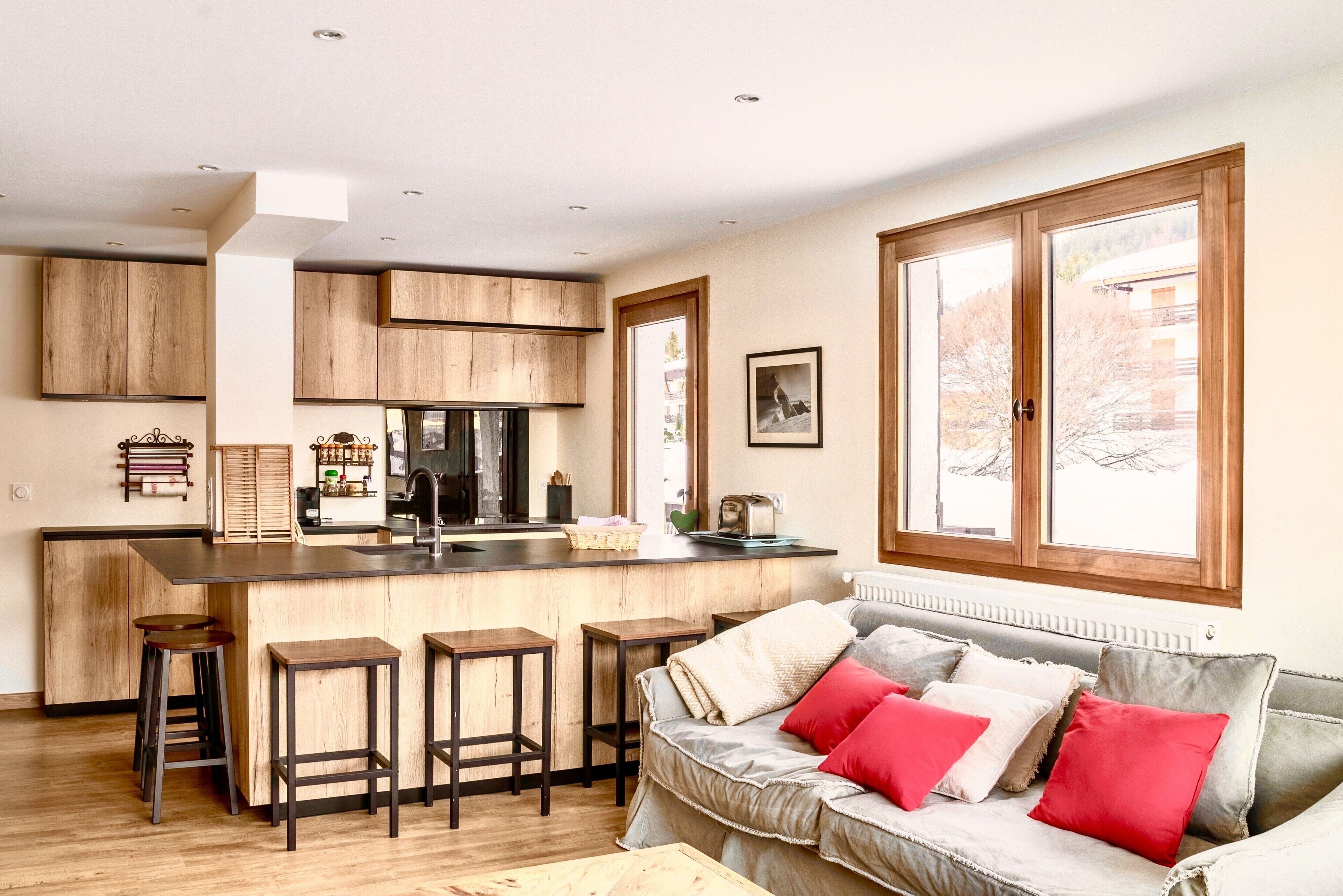 Een moderne keuken met houten kastjes, een centraal eiland met krukken, en een groot raam met uitzicht op de omgeving.