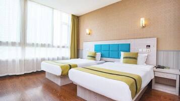 A hotel room with two beds, a large window with sheer curtains, a wooden floor, and a light-colored wall with a textured pattern.