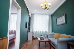 Una habitación con sofá, una mesita, televisor y ventana con cortinas.