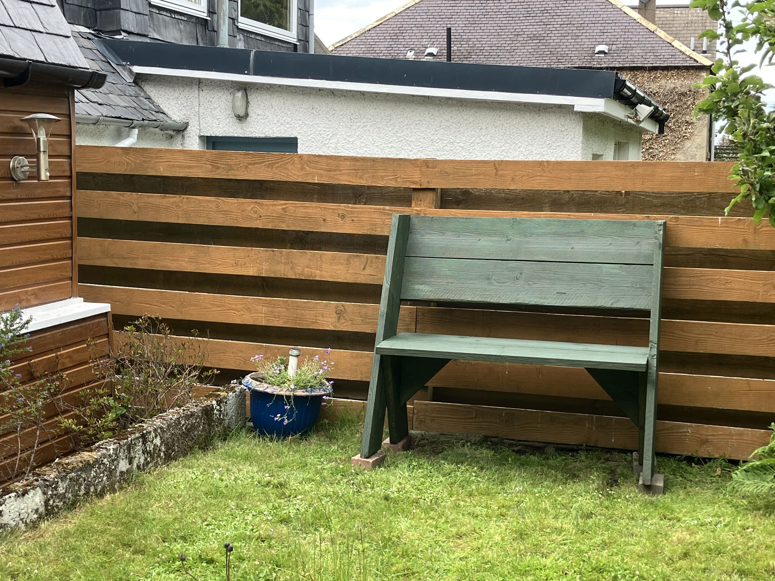 A wooden bench in a garden area with a white wall and a wooden fence in the background.