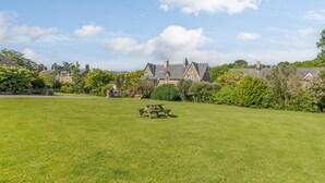 A well-maintained lawn with a picnic table in the foreground, surrounded by lush greenery and a stone building in the background. - Barnacre (Morpeth)