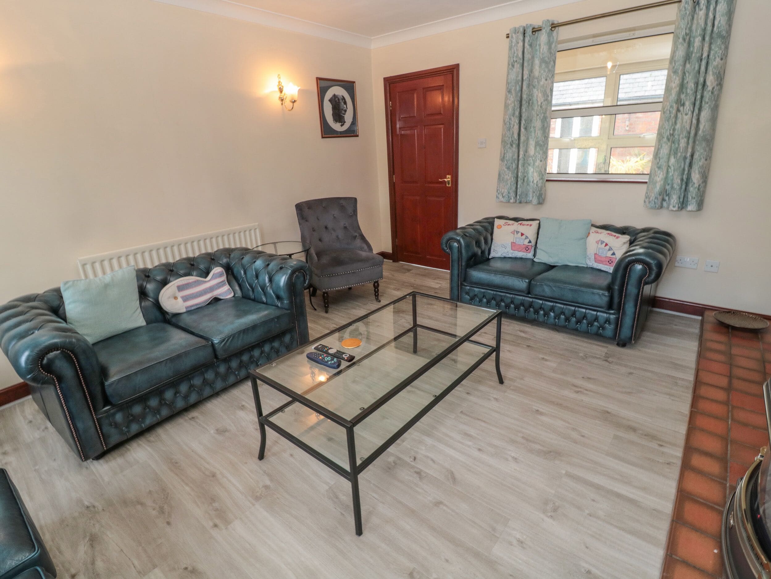 Een woonkamer met twee leren banken, een glazen salontafel, een houten vloer, een radiator, een deur en een raam met gordijnen.