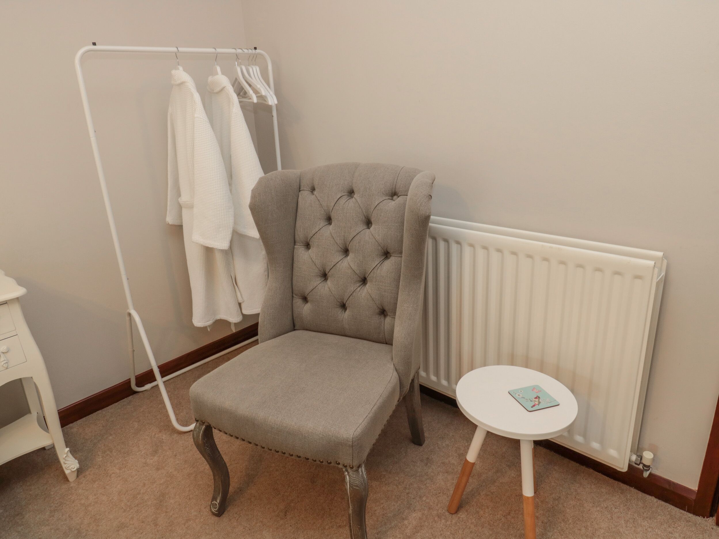 Ein Zimmer mit einem gepolsterten Sessel, einem Heizkörper, einem weißen runden Tisch und einem Kleiderständer mit bekleideten Kleidungsstücken.