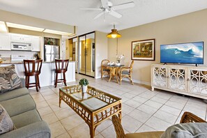 A modern living room with a sofa, a glass-top coffee table, and a dining area with wicker chairs. - Hawaiian decor, private lanai with tropical views (Lahaina)