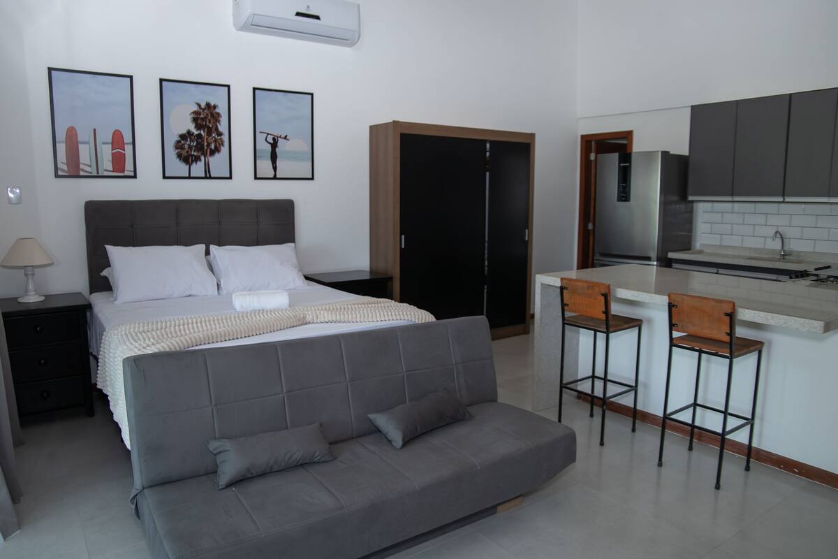 A modern bedroom with a large bed, a grey sofa, a small kitchen area with a fridge, and a bar stool.