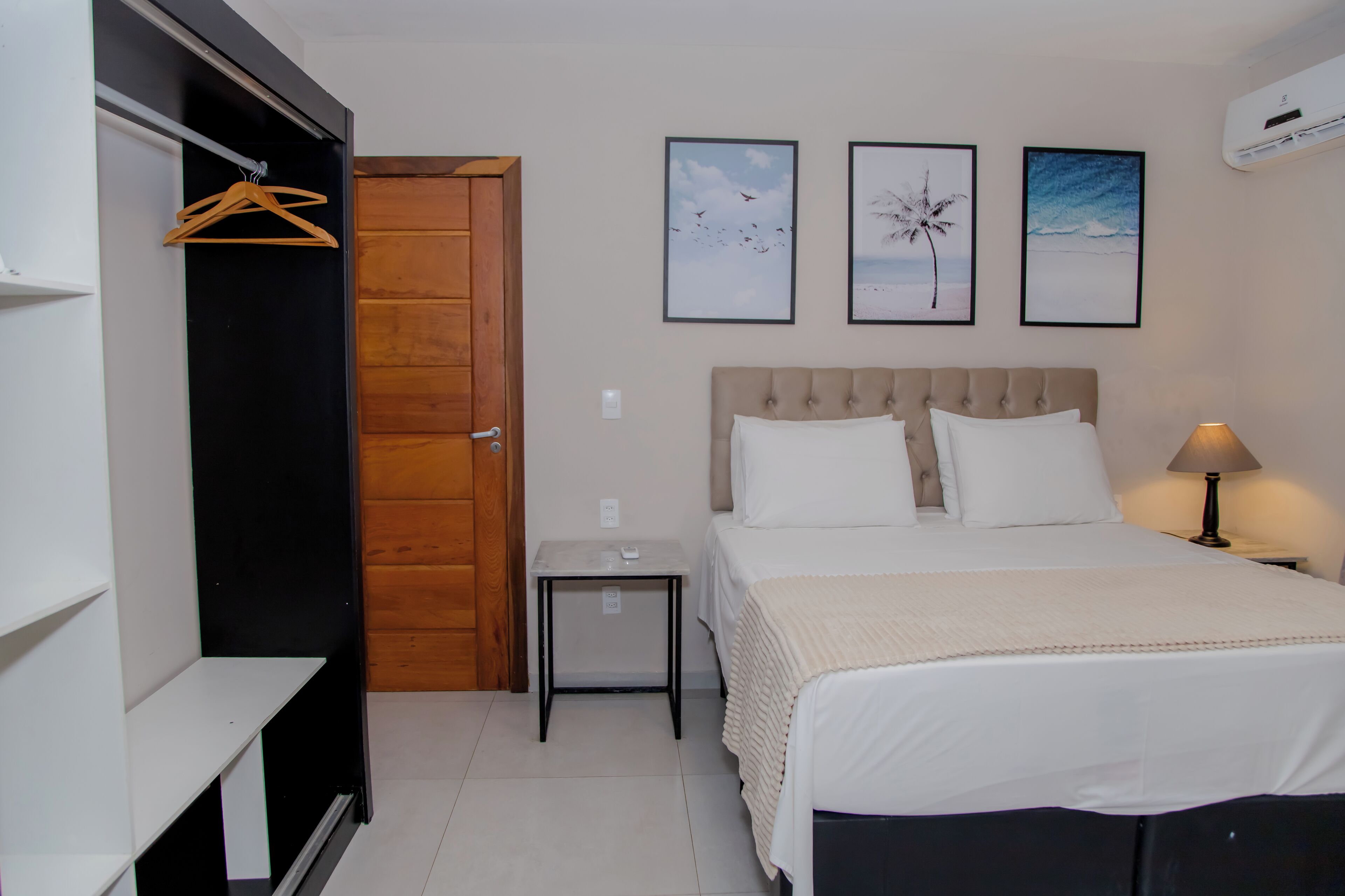 A bedroom with a bed, a bedside table, a nightstand, a hanging coat rack, and three framed pictures on the wall.