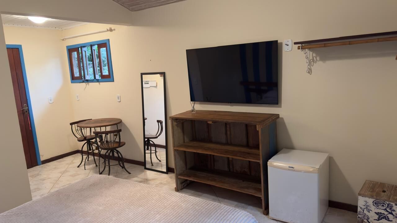 Una habitación con una mesa de comedor y sillas, un televisor montado en la pared, un refrigerador pequeño y una estantería de madera.