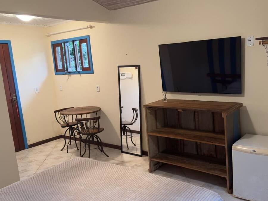 Una habitación con una mesa de comedor y sillas, un televisor montado en la pared, un refrigerador pequeño y una estantería de madera.