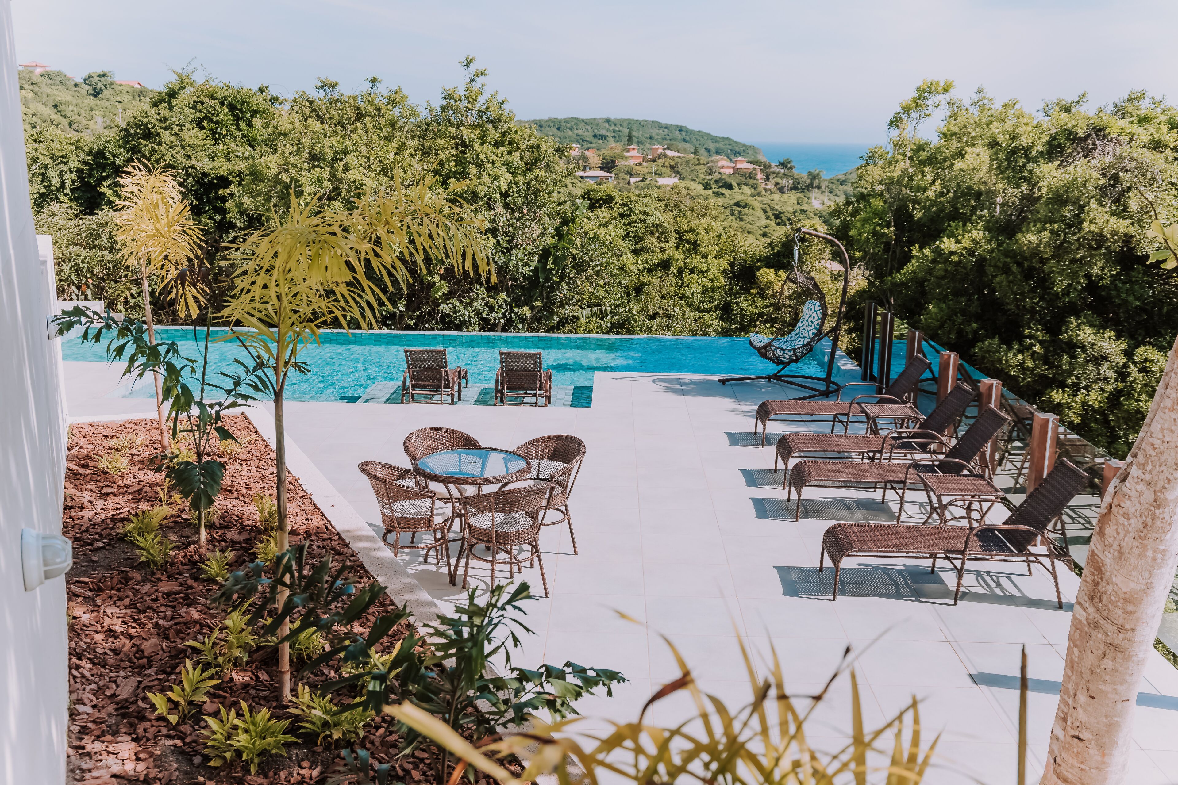 An outdoor pool area with lounge chairs and a table, surrounded by tropical plants and overlooking a scenic view.