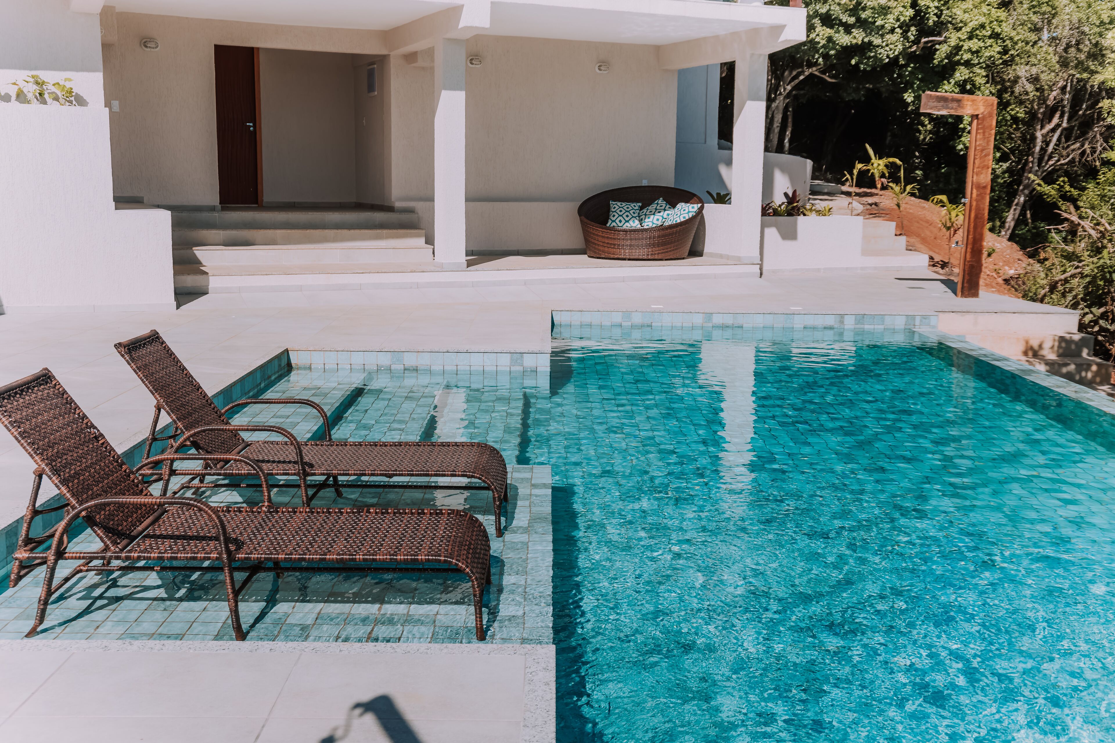 A swimming pool with lounge chairs and a wicker chair, surrounded by a white building and greenery.