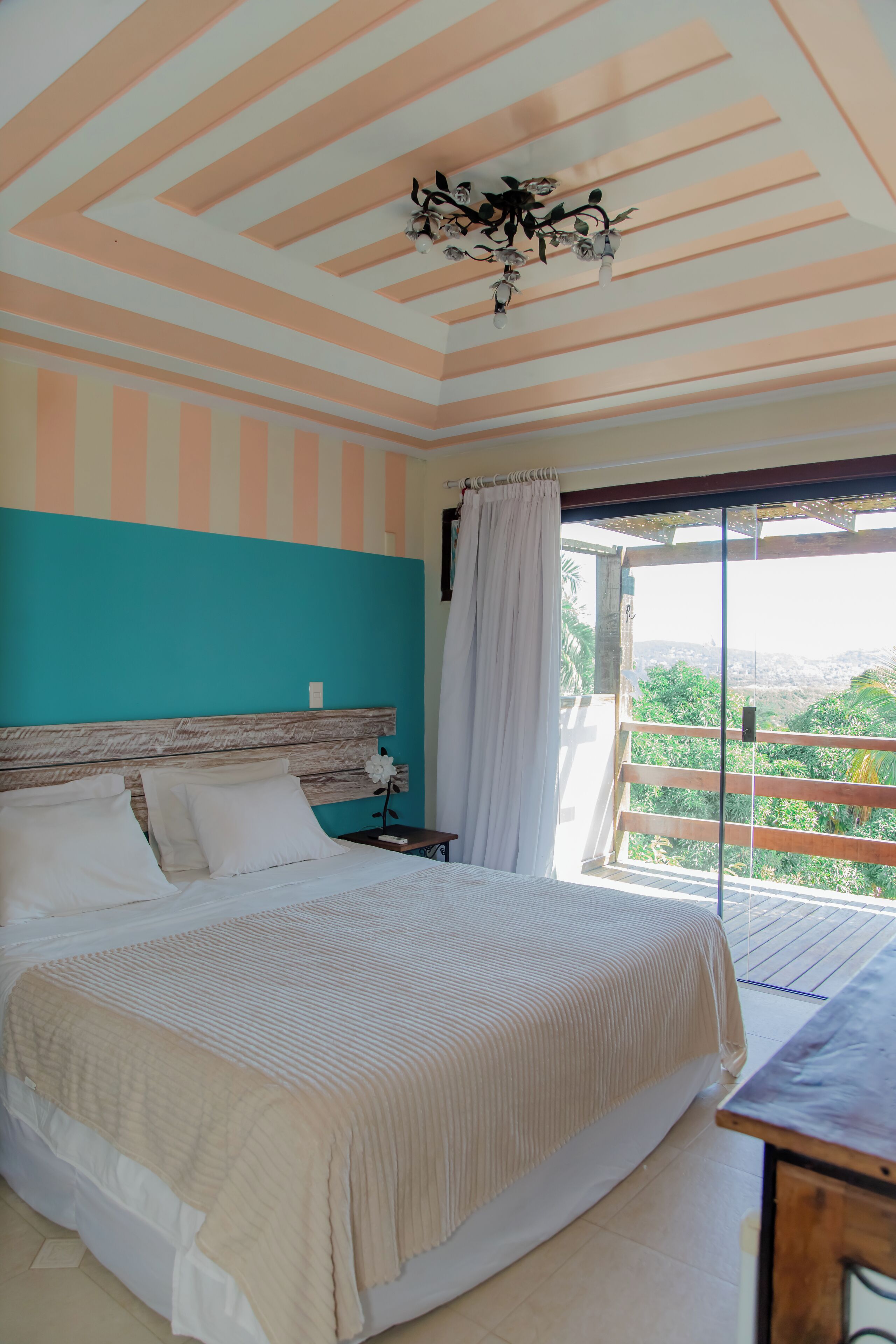 A bedroom with a bed, a nightstand, a chandelier, and a view of greenery through a balcony door.