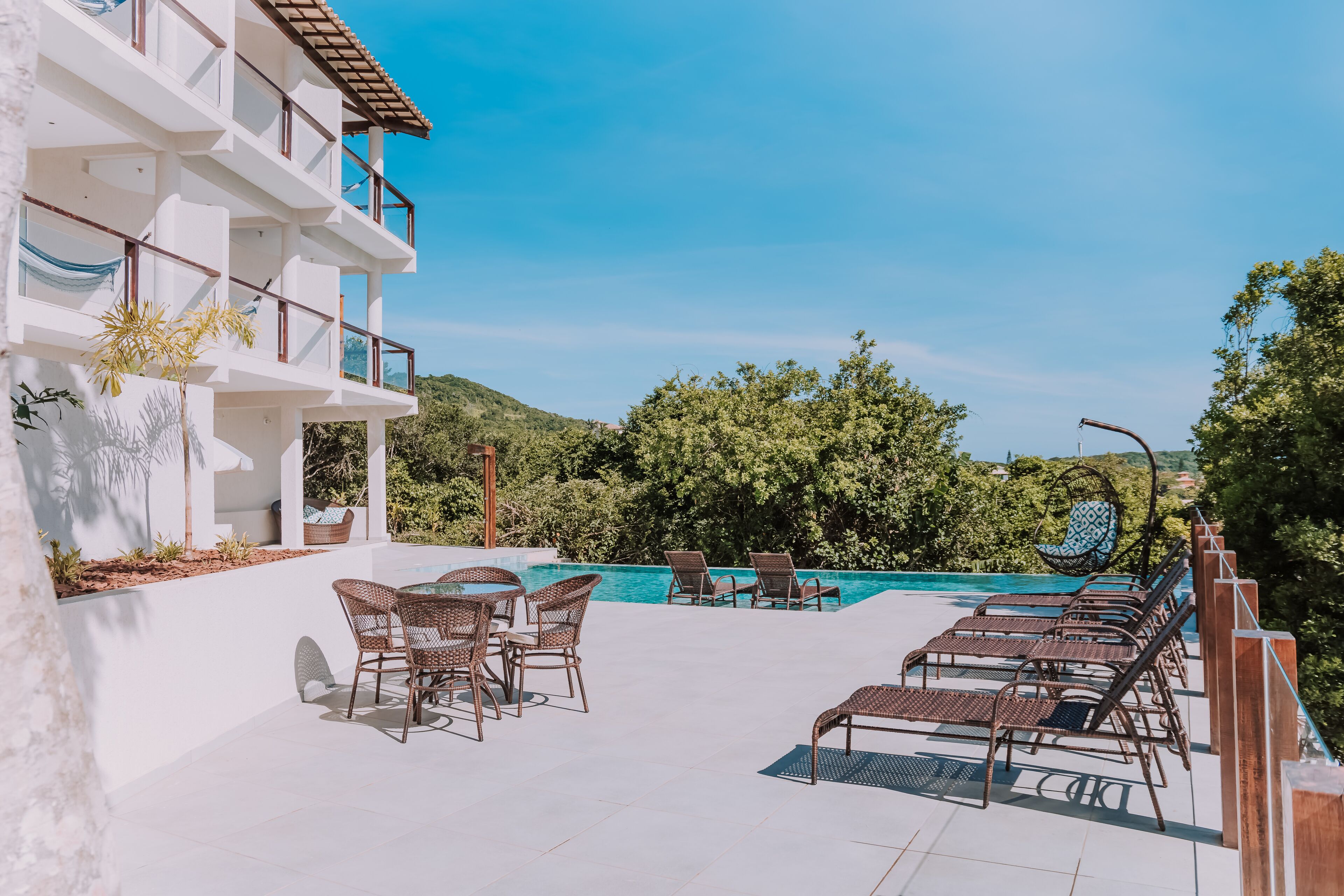 Un espace aménagé au bord de la piscine, avec des chaises longues et une table à manger, entouré de verdure et surplombé d’un ciel bleu azur.