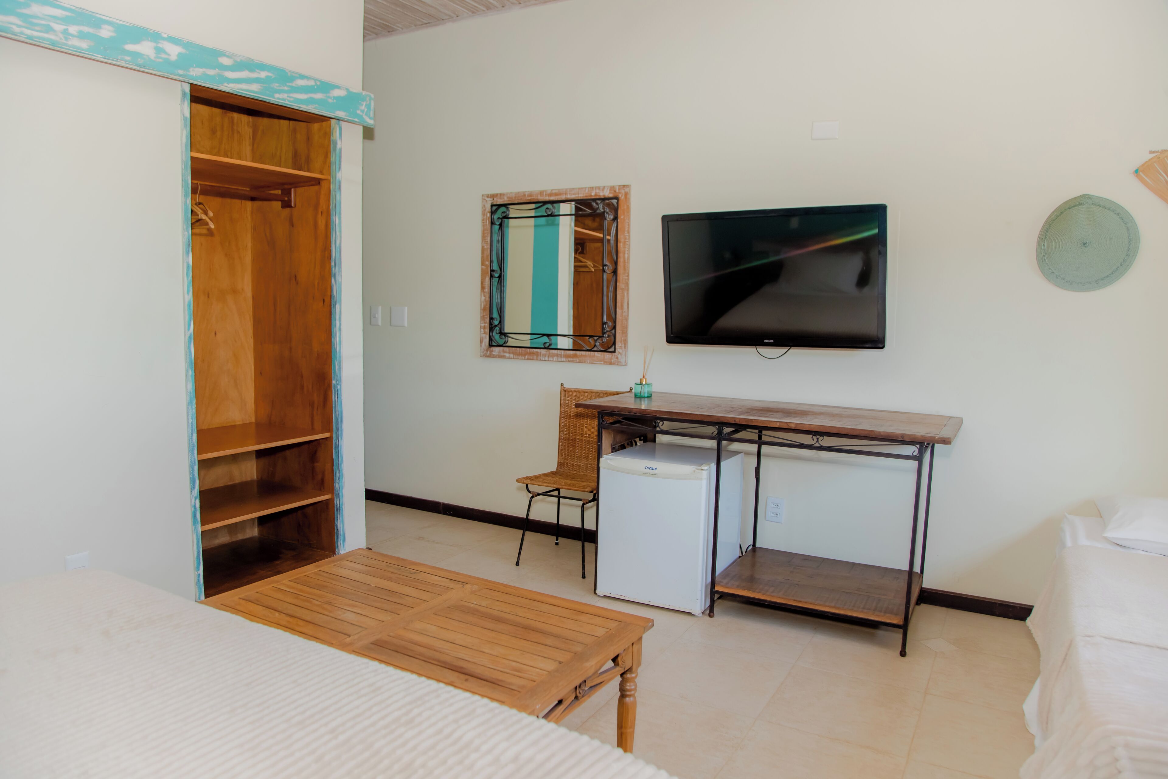 A room with a wooden wardrobe, a small desk with a chair, a television mounted on the wall, and a bed with white bedding.