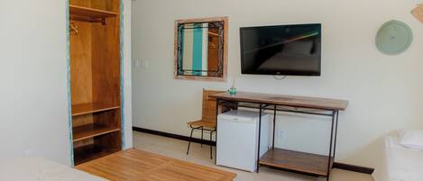 A room with a wooden wardrobe, a small desk with a chair, a television mounted on the wall, and a bed with white bedding.