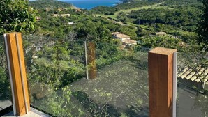 Una terraza con barandilla de vidrio que domina un paisaje exuberante con colinas y una vista distante del mar.