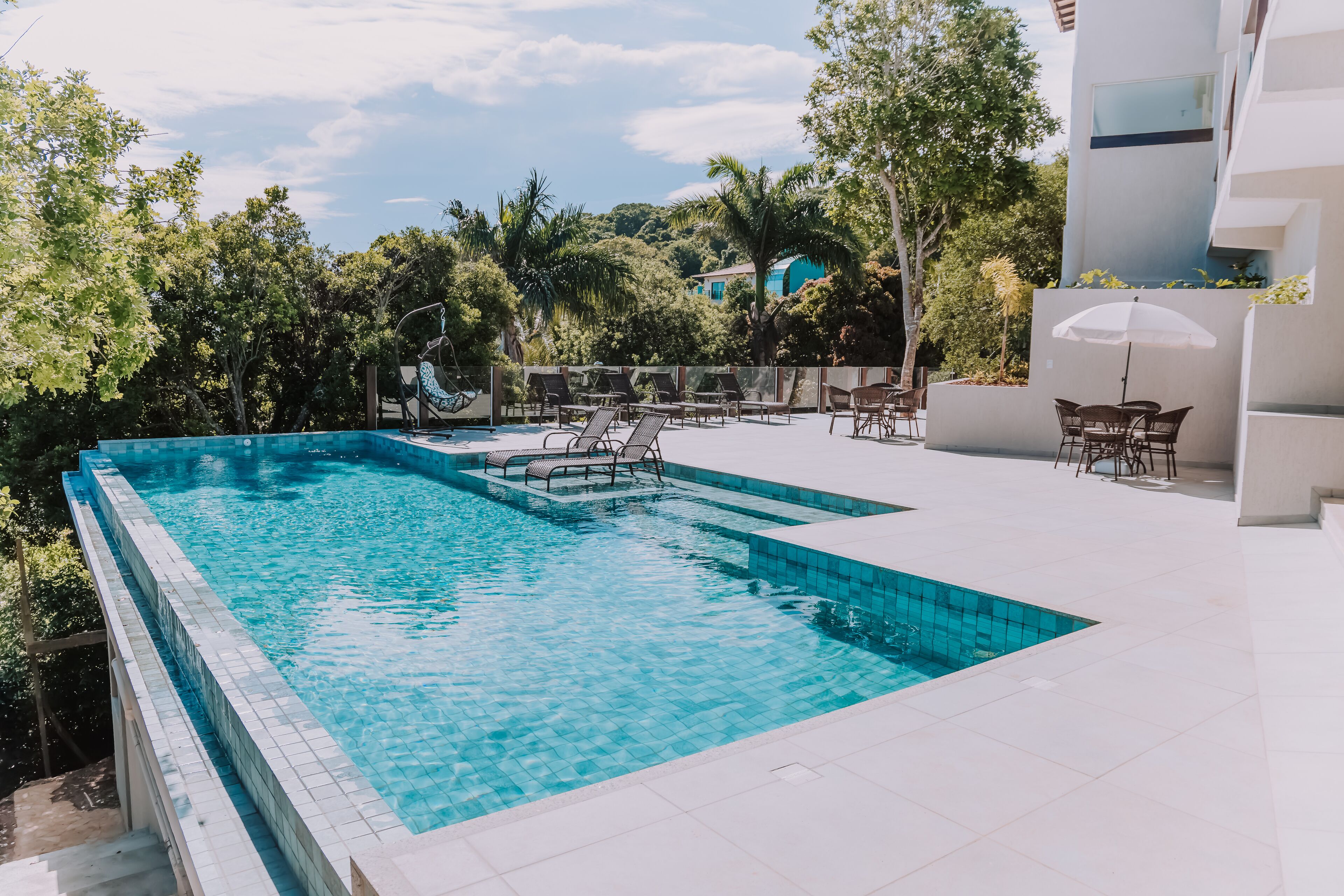 A rectangular swimming pool with clear blue water, surrounded by white tiled deck, and furnished with multiple lounge chairs and a table with a parasol.
