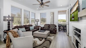 A modern living room with a sectional sofa, a coffee table, and a ceiling fan. There are large windows with blinds, a TV mounted on the wall, and a dining area visible through an open door.